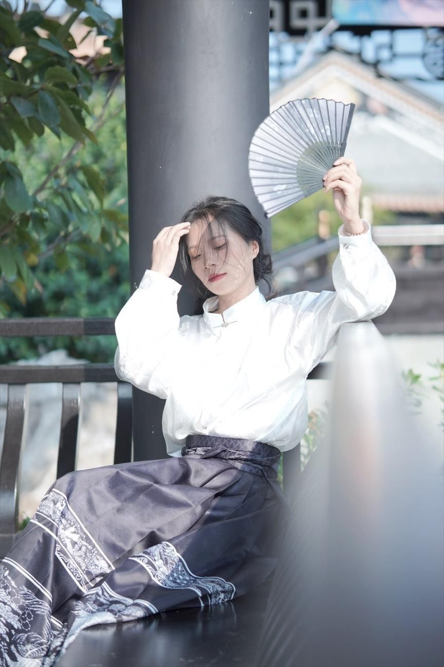 Woman in traditional Hanfu outfit holding a fan, seated on a bench in a serene outdoor setting.