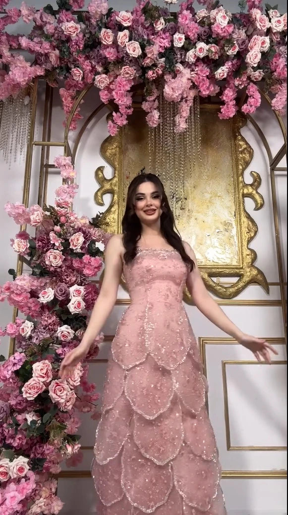Model in a customized pink beaded engagement dress with tiered skirt, surrounded by floral decor.
