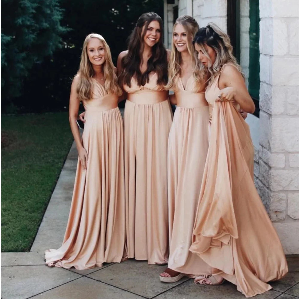 Four bridesmaids in floor-length V-neck satin dresses in soft peach color, posing outdoors with greenery.
