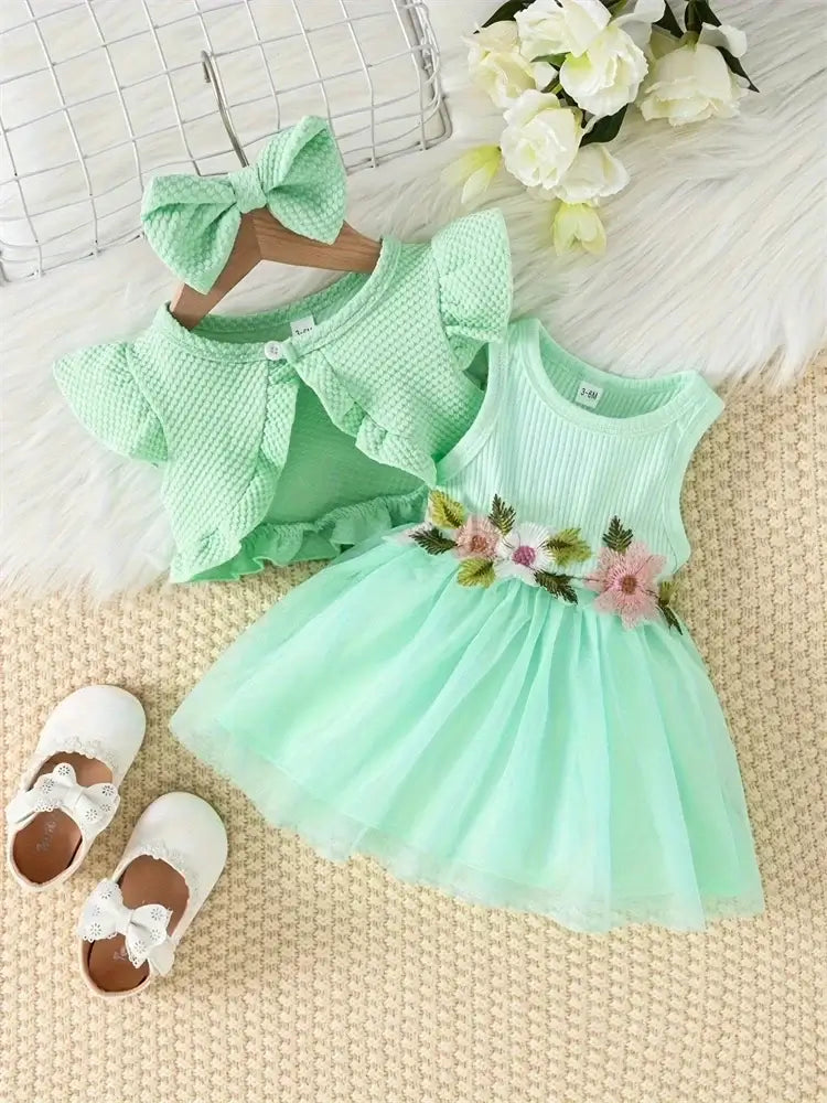 Newborn baby girls dress set in mint green with floral details, matching cardigan, and bow headband on a textured surface.