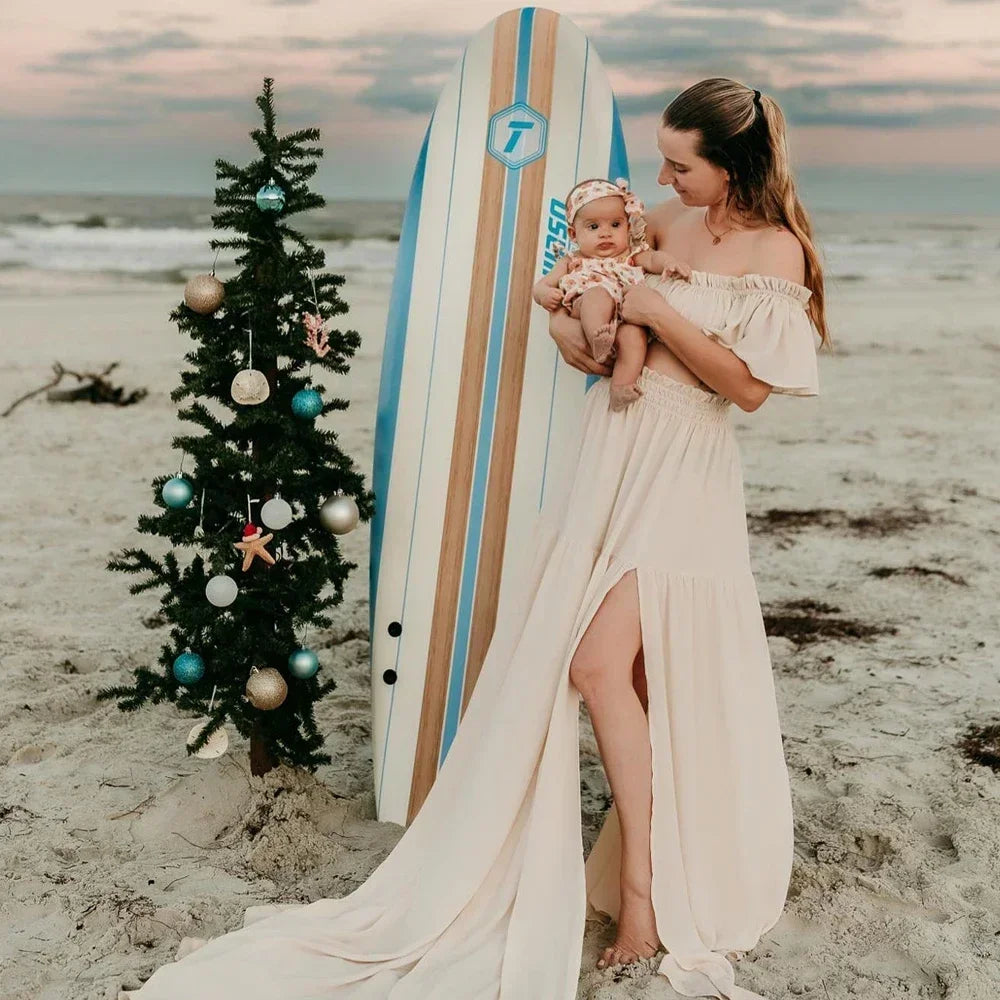 A mother in a flowy off-shoulder maternity dress holding a baby by the beach near a surfboard and a decorated Christmas tree.