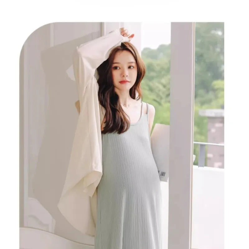 A pregnant woman in a sleeveless maternity dress, wearing a light jacket, standing by a sunny window.