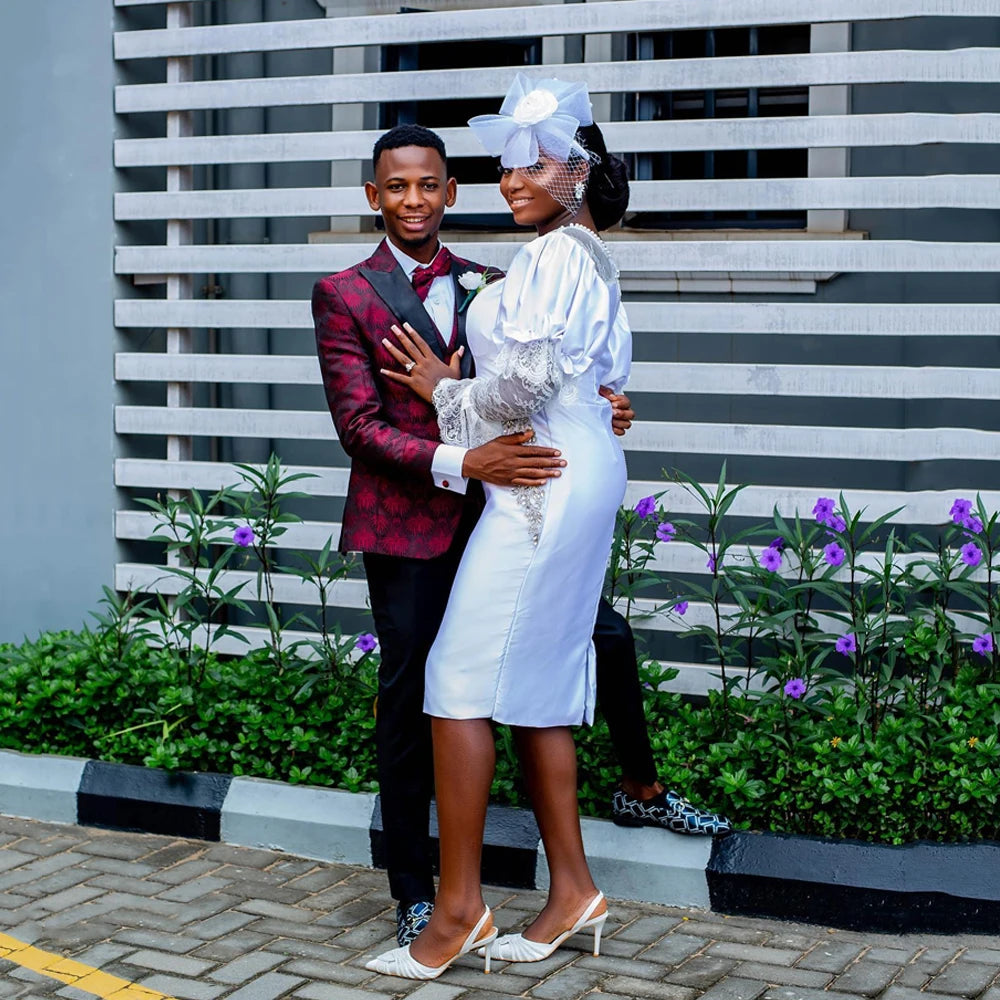Stylish couple in formal attire, embracing in front of a modern backdrop with purple flowers.