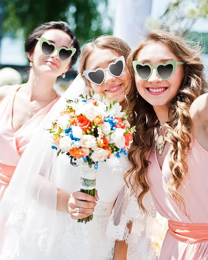 Three smiling bridesmaids wearing heart-shaped sunglasses, one holding a bouquet, at a summer wedding.
