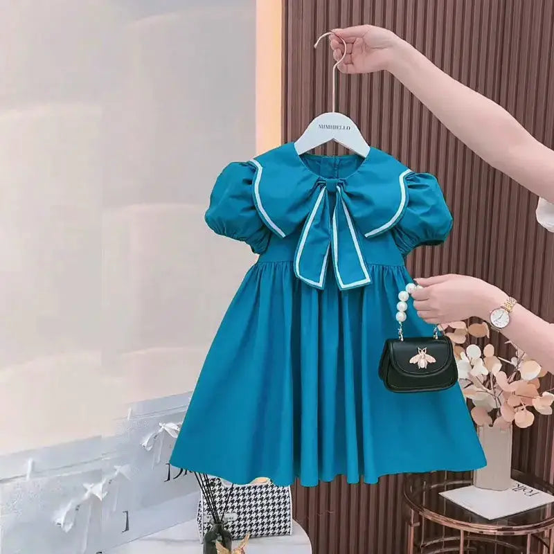 Cute summer blue dress for baby girls with puff sleeves and a Peter Pan collar, paired with a small handbag.