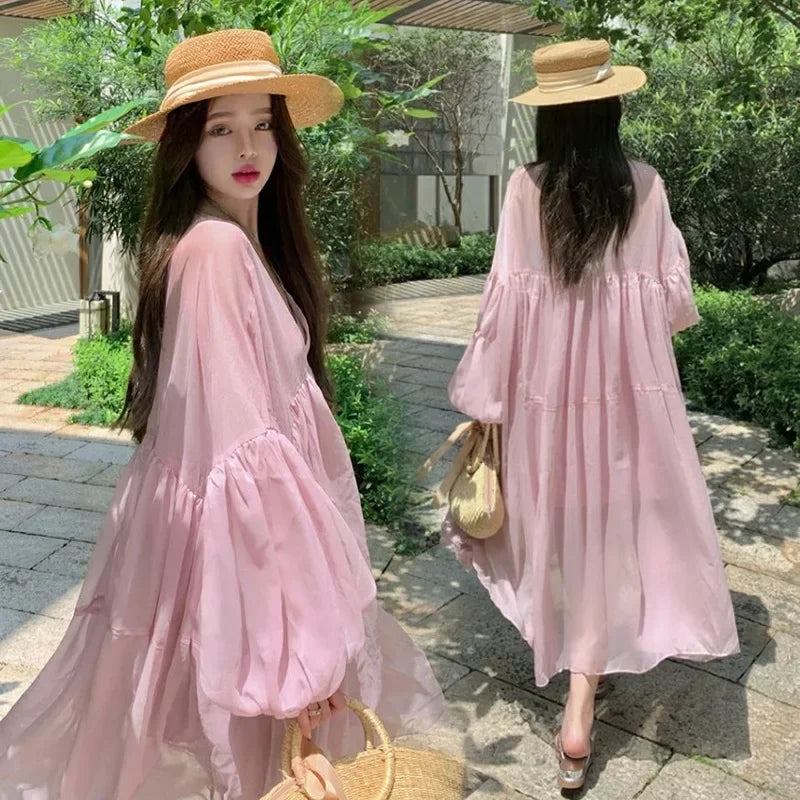 Woman in pink chiffon maternity dress with puff sleeves, walking outdoors in a summer setting, wearing a straw hat.