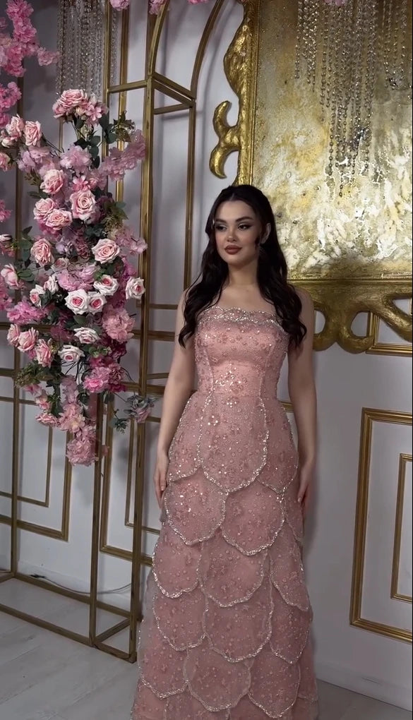 Elegant woman in a pink beaded engagement gown with a tiered skirt, posed in front of floral decor.