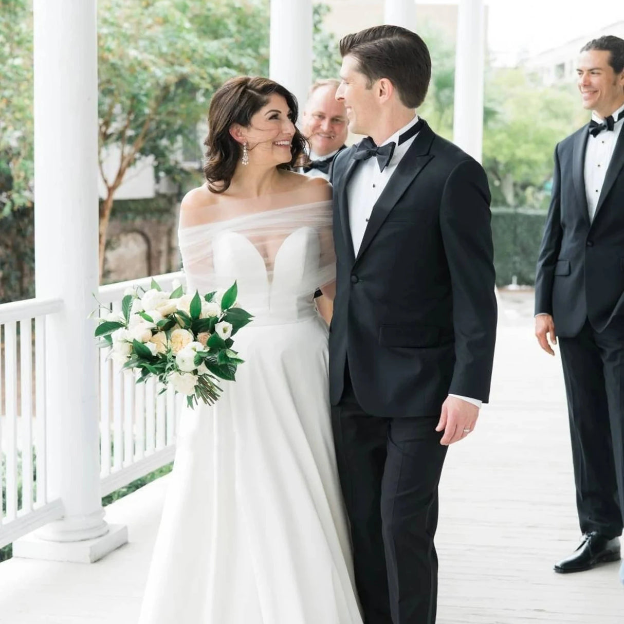 Bride wearing a sheer wedding jacket over her gown, smiling at her groom while holding a bouquet.