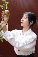 Woman in a white blouse reaching for a fruit hanging from a branch in a serene outdoor setting.