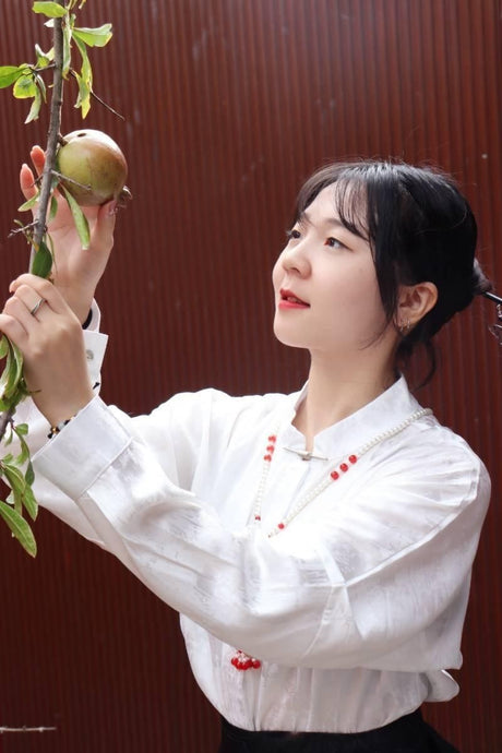 Woman in a white blouse reaching for a fruit hanging from a branch in a serene outdoor setting.