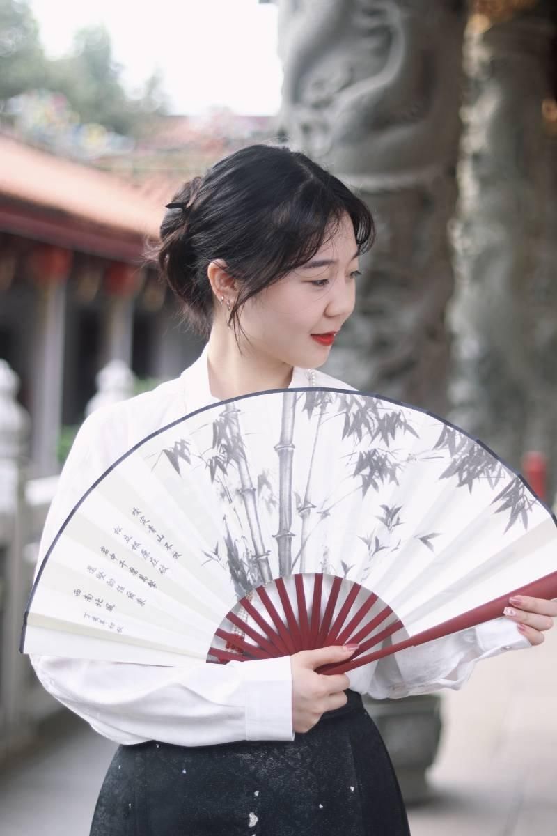 Woman holding a traditional bamboo fan, wearing a white blouse, showcasing a serene expression in a beautiful outdoor setting.
