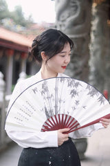 Woman holding a traditional bamboo fan, wearing a white blouse, showcasing a serene expression in a beautiful outdoor setting.