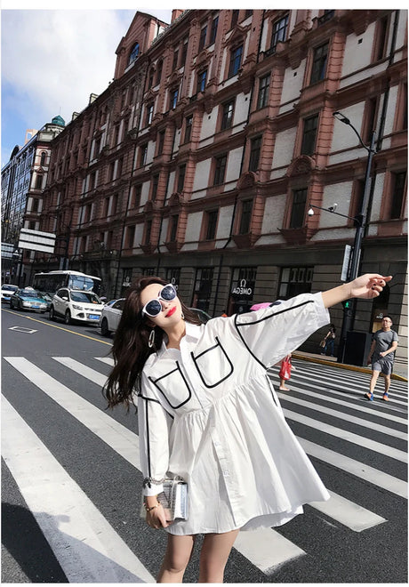Model wearing a solid color maternity shirt dress with pockets, enjoying a sunny day on a city street.