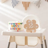 Wooden baby milestone flower shape displaying '2 Months' next to a colorful birthday cake on a high chair.
