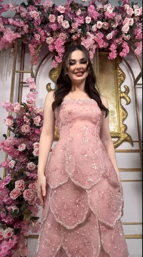 Model wearing a pink beaded engagement gown with a tiered skirt, surrounded by floral decorations.