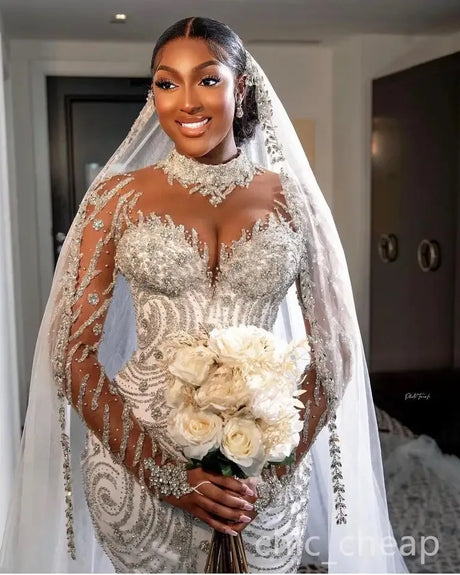 Bride in a luxurious beaded mermaid wedding dress with sheer sleeves, holding a bouquet of white flowers.