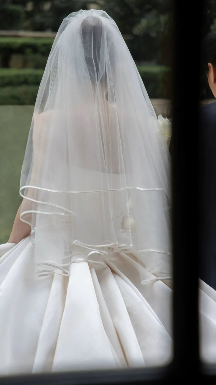 Bride in elegant satin wedding dress and veil viewed from behind, showcasing classic bridal style and lace details.