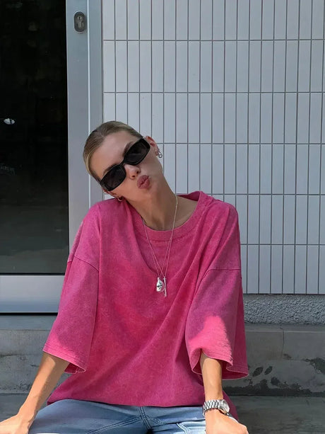 Woman in pink casual t-shirt posing playfully, wearing sunglasses and sitting outdoors.
