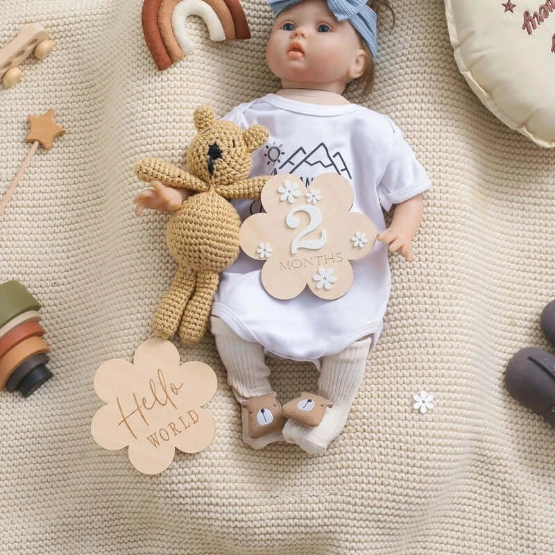 Wooden baby milestone set with a doll, featuring 'Hello World' and '2 Months' signs on a cozy blanket.