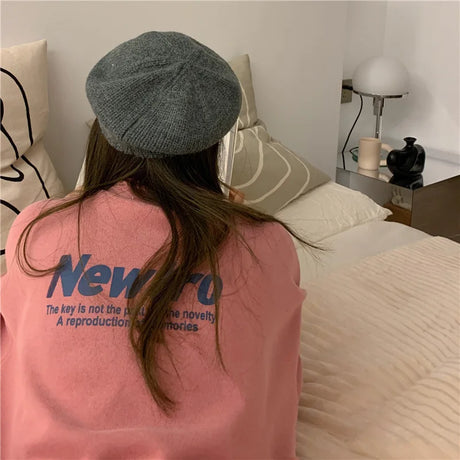 Young woman in a pink oversized tee and gray beret sitting on a bed with stylish decor.