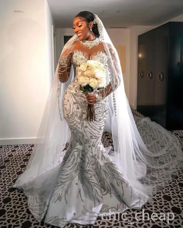 Luxury beaded mermaid wedding dress with sheer sleeves, model holding flowers in an elegant setting.