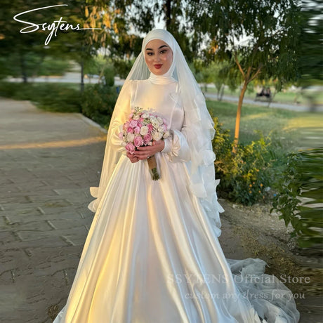 Muslim A-Line satin wedding dress with high collar, model holding bouquet in a scenic outdoor setting.