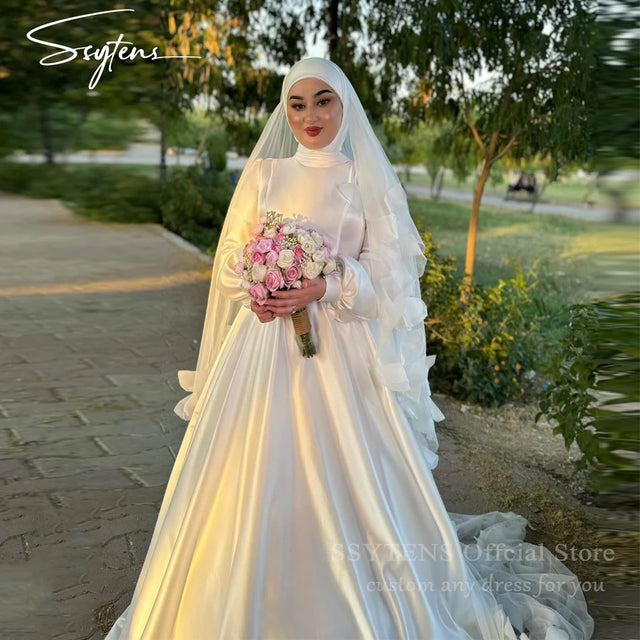 Muslim A-Line satin wedding dress with high collar, model holding bouquet in a scenic outdoor setting.