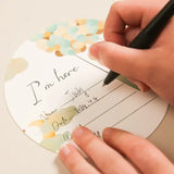 Person writing on a circular baby milestone card with a decorative design and text "I'm here".