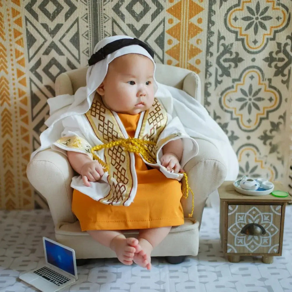 Adorable baby dressed in a traditional outfit, seated in a cozy chair with a decorative background.