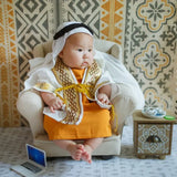 Adorable baby dressed in a traditional outfit, seated in a cozy chair with a decorative background.