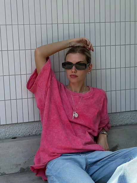 Woman wearing a pink casual t-shirt with loose fit and sunglasses, sitting against tiled background.