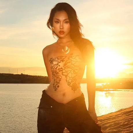 Model posing against a sunset backdrop near water, showcasing body art and stylish jewelry.
