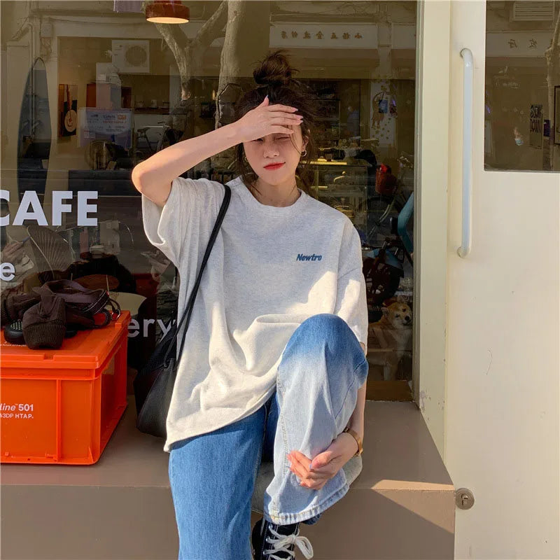 Casual oversized Harajuku streetwear tee on a young woman in a café, paired with loose jeans and a shoulder bag.