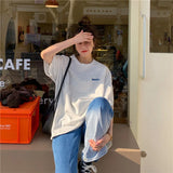 Casual oversized Harajuku streetwear tee on a young woman in a café, paired with loose jeans and a shoulder bag.
