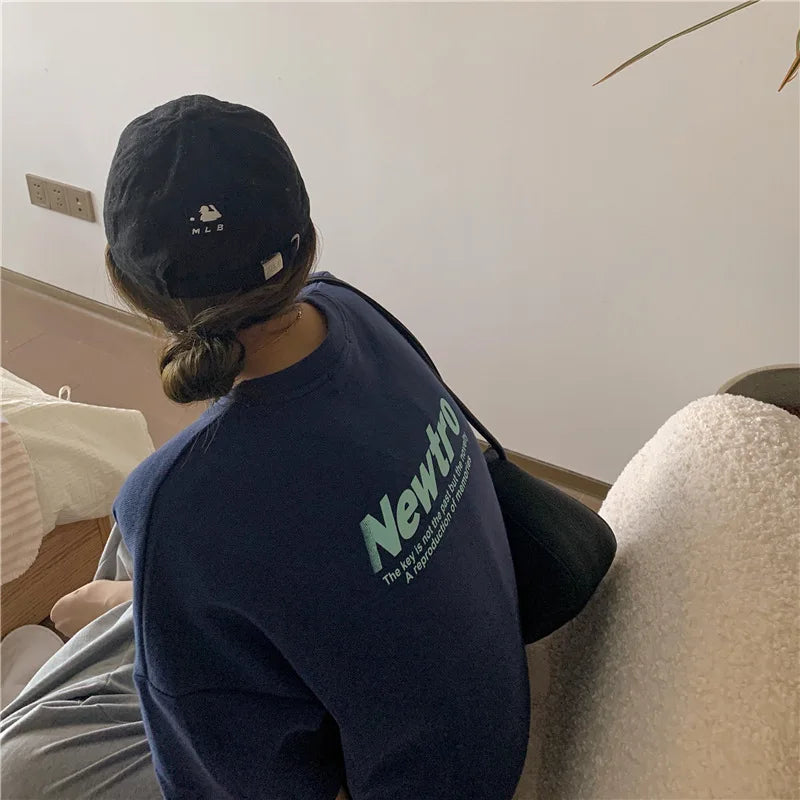 Woman wearing a navy oversized sweater with 'Newtro' print and a black cap, seated in a cozy room setting.