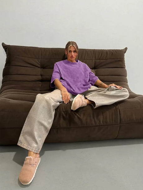Woman wearing a purple oversized casual t-shirt and silver pants, sitting on a brown couch in a modern setting.