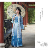 Elegant woman in a blue Hanfu cosplay dress holding a decorative parasol in a scenic outdoor setting.