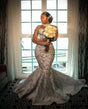 Elegant woman in a luxury beaded mermaid wedding dress with sheer sleeves, holding a bouquet in front of a window.