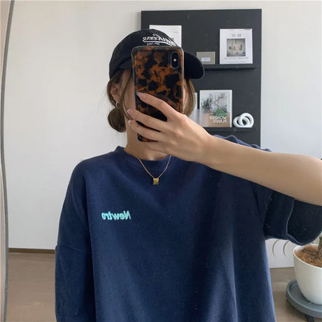A young woman wearing an oversized navy Harajuku streetwear tee with the text 'Newton' reflected in a mirror.