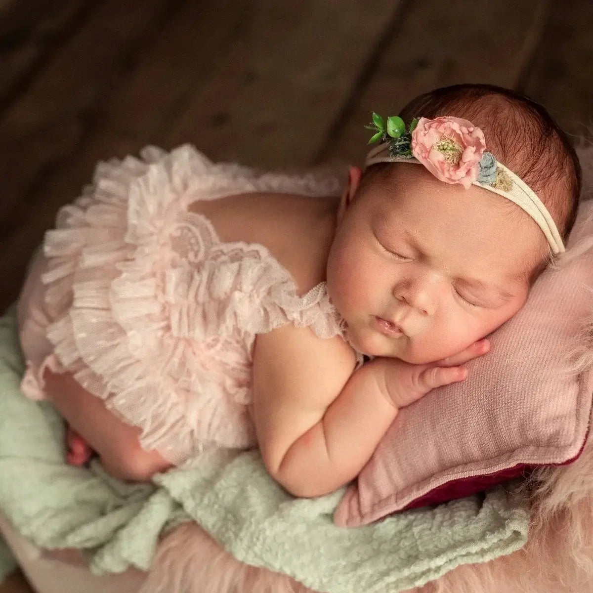 Newborn baby girl sleeping in pink lace outfit with floral headband, resting on soft cushions.