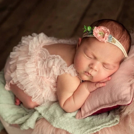 Newborn baby girl sleeping in pink lace outfit with floral headband, resting on soft cushions.