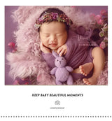 Newborn girl wearing a faux flower headband, smiling with a purple backdrop and soft textures, perfect for baby photography.