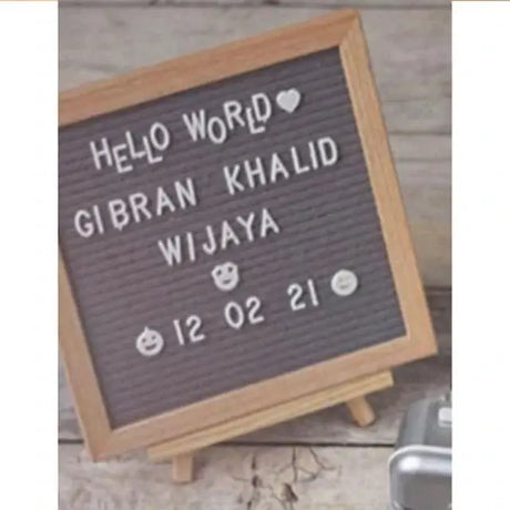 Letter board displaying "Hello World Gibran Khalid Wijaya 12 02 21" with decorative emojis on a wooden background.