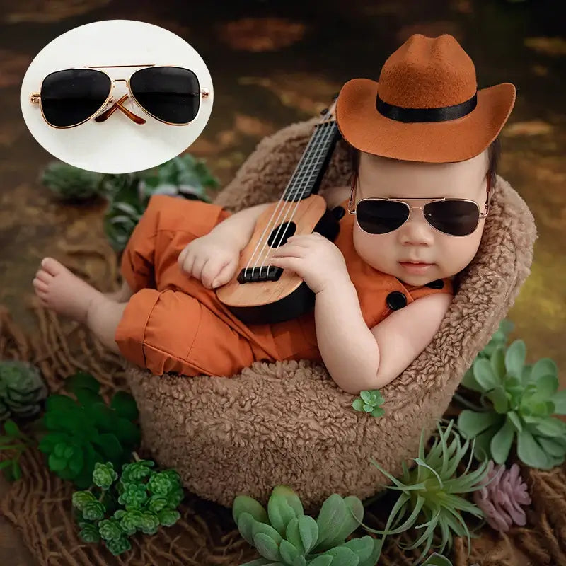 Newborn wearing round sunglasses and a brown hat, posing with a mini guitar, showcasing baby glasses photography accessories.