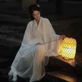 Woman in traditional white Hanfu dress sitting beside a glowing lantern in the night.