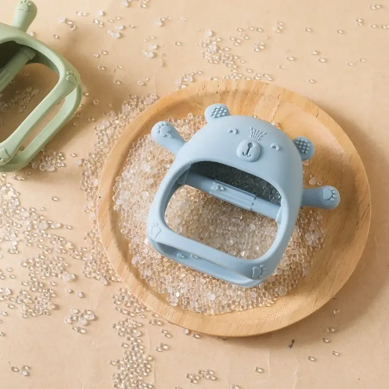 Soft blue bear-shaped baby teether glove on a wooden plate surrounded by decorative beads.