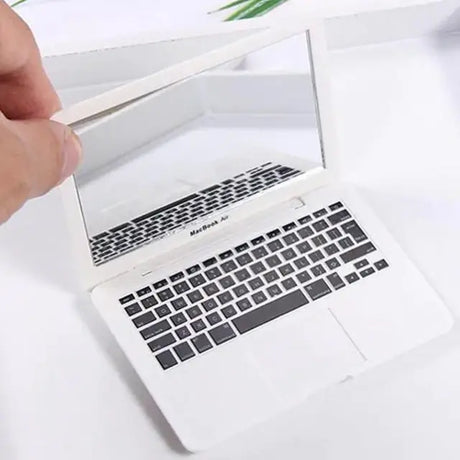 Hand holding a small white laptop mirror resembling a MacBook Air, featuring a realistic keyboard design.