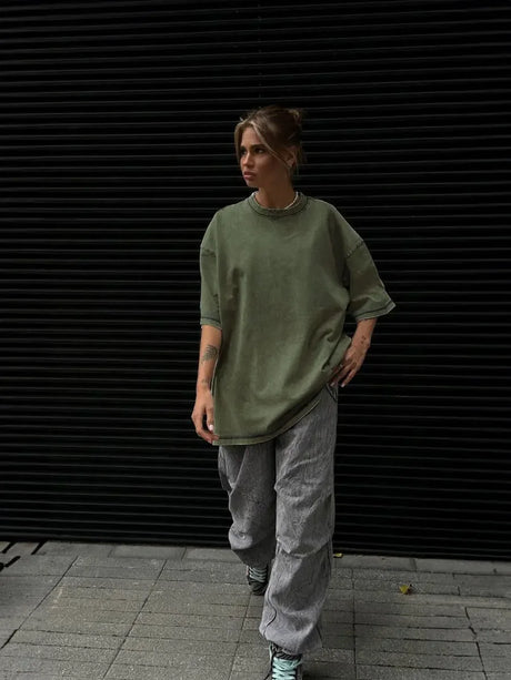 Woman in oversized olive green t-shirt and gray trousers walking against a black background.