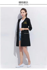 Model wearing a black salon beauty lab coat, showcasing pockets and stylish design, perfect for beauty professionals.