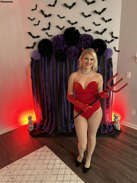 Woman in red velvet strapless bodysuit with devil horns, posing in front of Halloween-themed backdrop.